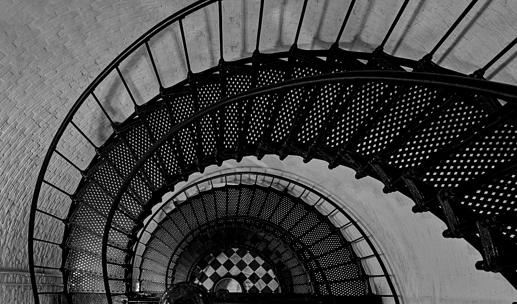 An iron spiral staircase descends to a chequred floor inside a lighthouse.